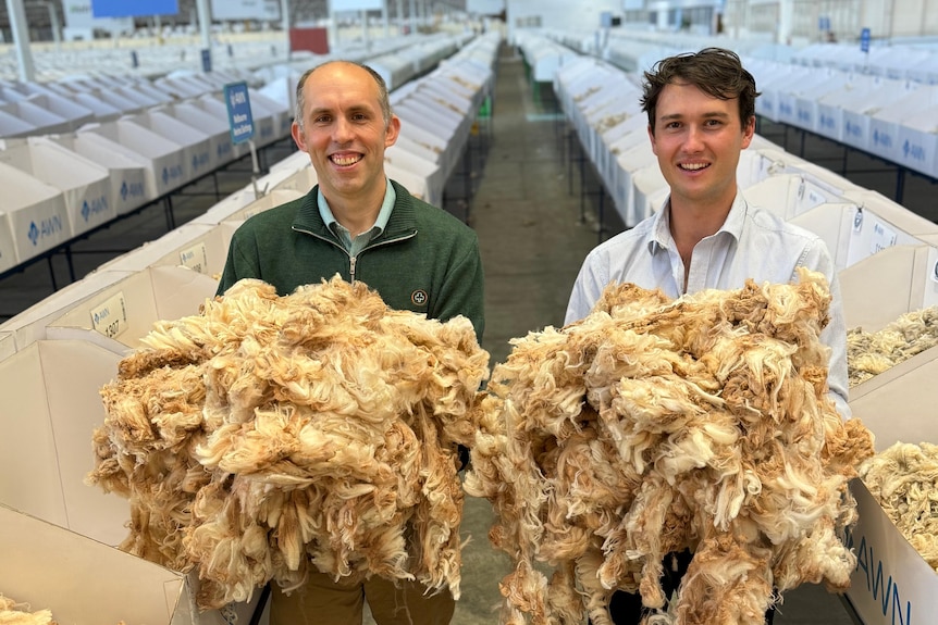 Two men stand holding sheep's wool in their hands.