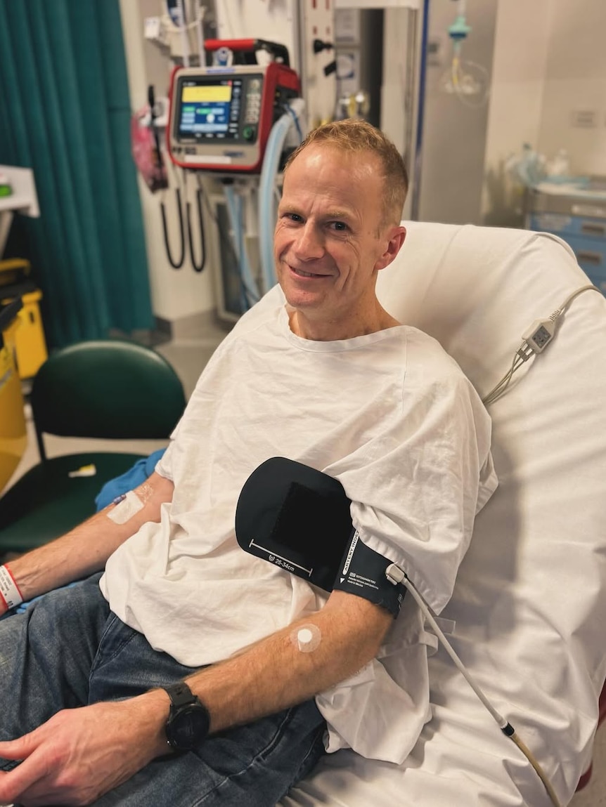 Man wears a white t-shirt seated in hospital room. His forearm is wrapped in blood pressure monitor
