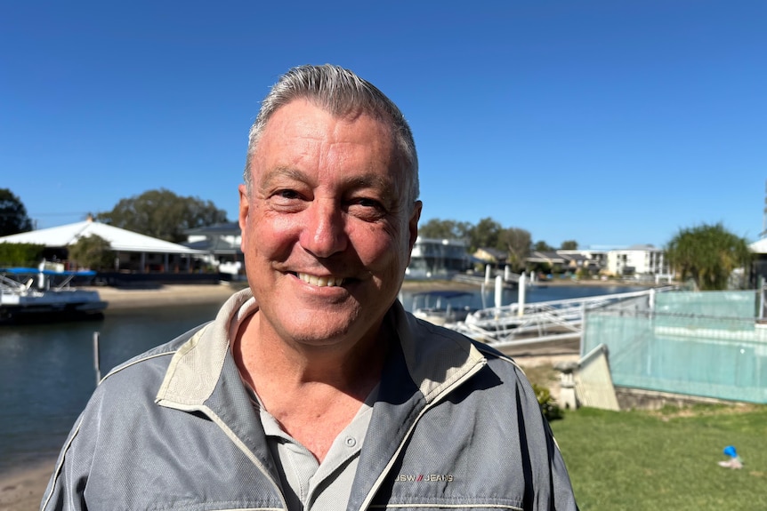 A smiling, midddle-aged man with short grey hair stands near a waterway.