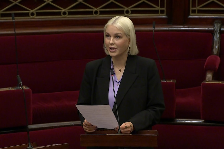 A woman with chin length blonde hair in a dark jacket and purple shirt stands in her parliament seat holding papers.