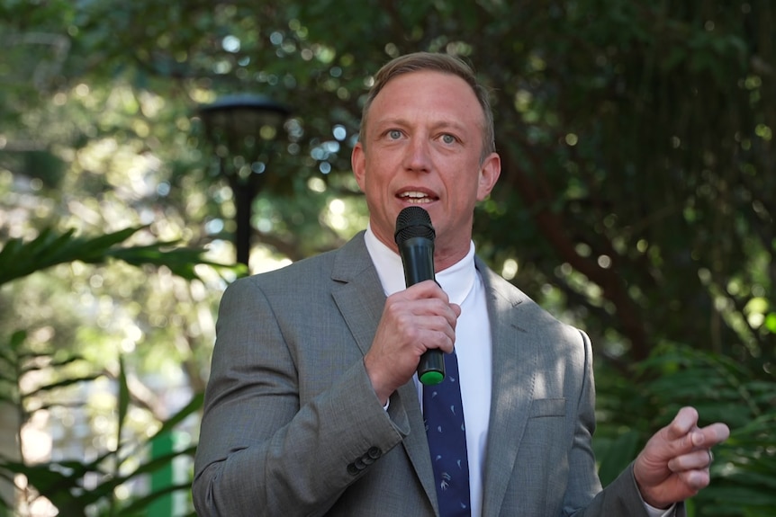 A man in a grey suit speaking into a microphone outside.