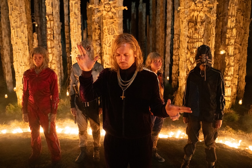 A group of people in tracksuits stand inside a ring of fire in front of bone towers
