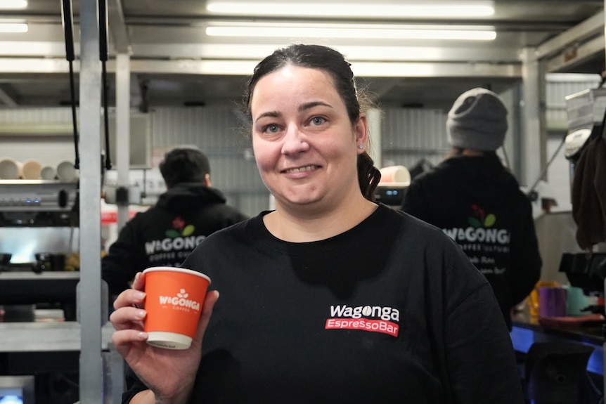A woman with dark hair pulled into a ponytail holds up an orange paper coffee cup.