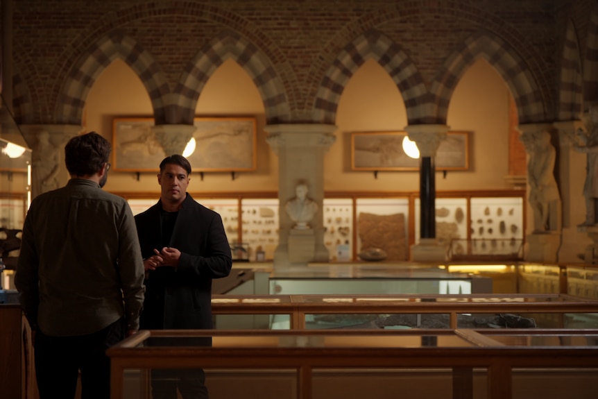 Marc stands in museum archives in dim lighting talking to an expert who has their back to us.