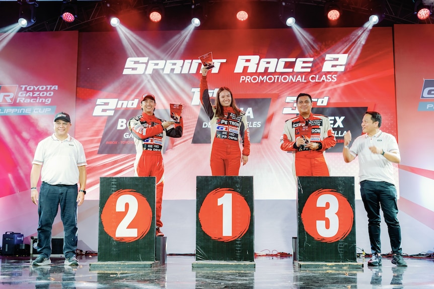 A woman stands on a podium in first spot while two men stand behind podiums 2 and 3.