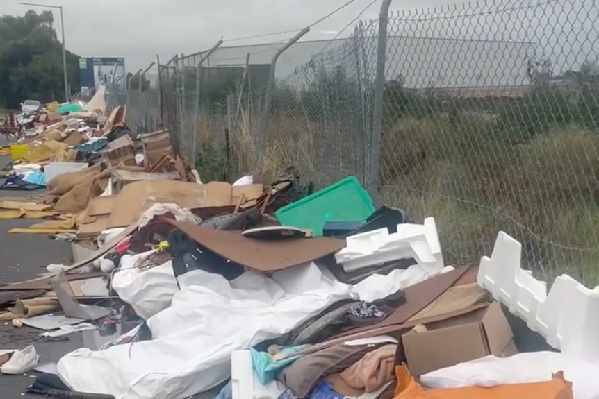 Piles of rubbish along a fence, in a carpark, alongside a road.