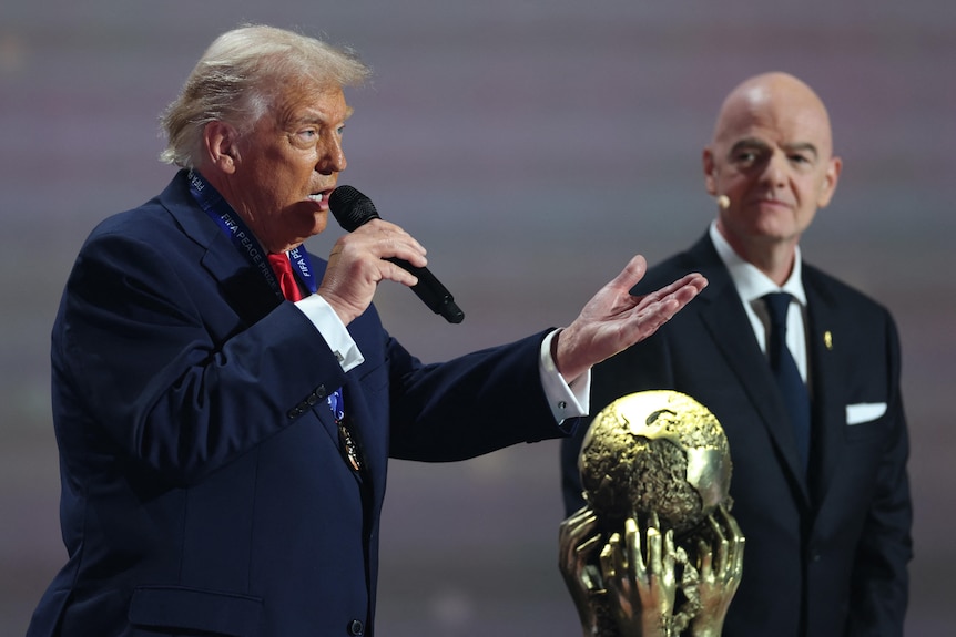 Donald Trump holds a microphone and speaks next to the World Cup trophy.