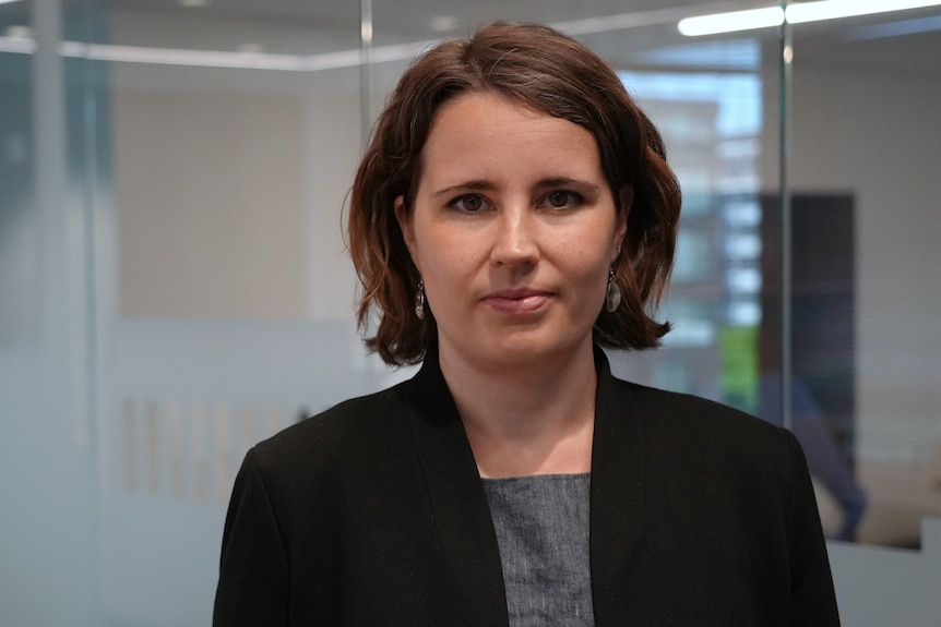 A woman with a brunette bob wearing a black blazer stands in a nondescript office with a neutral facial expression.