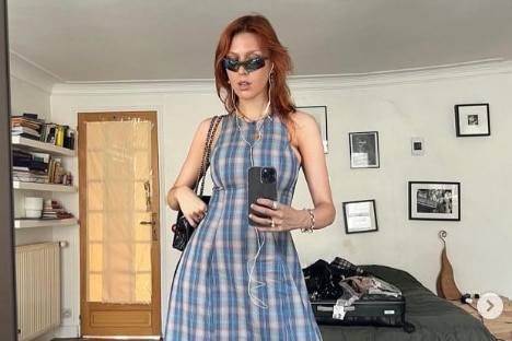 Young woman with red hair takes a photo in a mirror of herself wearing a sleeveless blue patterned dress.
