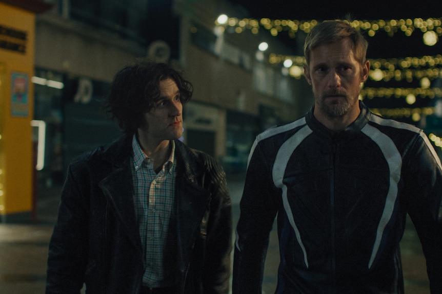 A brunette and a blond man walk next to each other through a dimly lit street