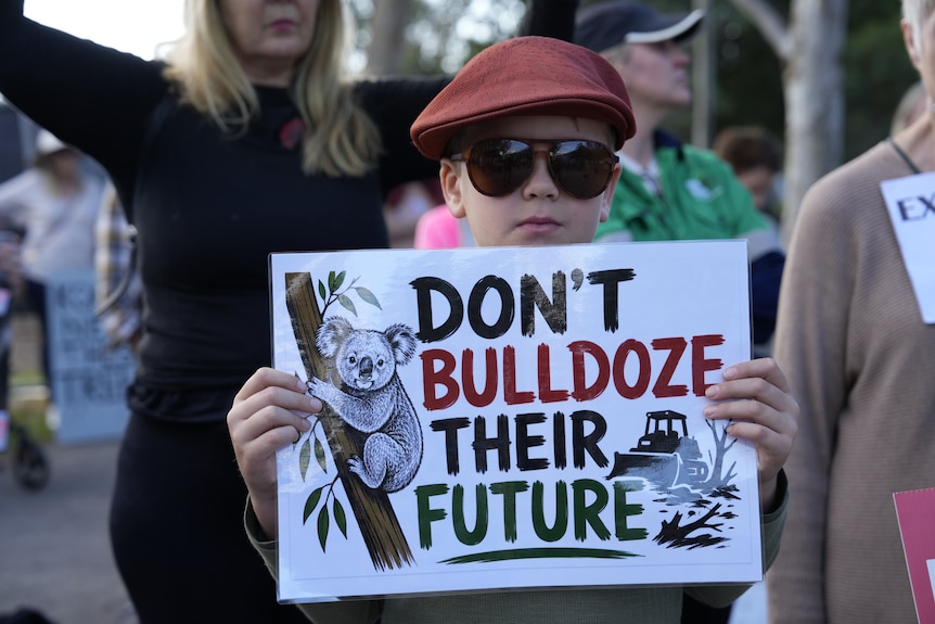 A young boy in sunglasses holds up a laminated sheet which reads: "don't bulldoze their future"