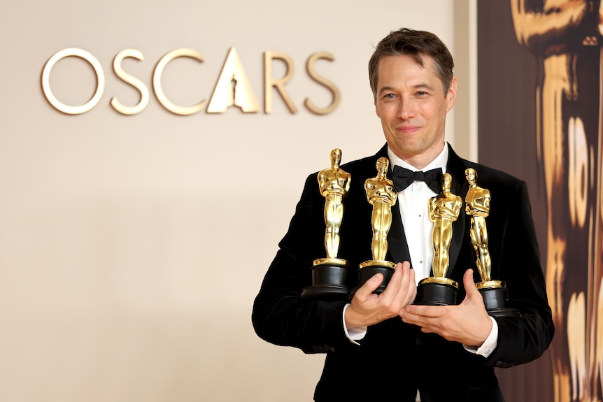 A man in a tuxedo holds four golden statues in front of a sign that says Oscars.