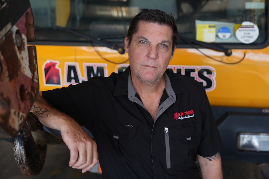White man with black short cut hair, wearing a black polo shirt, stading in front of a yellow vehicle, with a serious expression