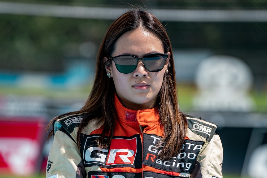 A woman in racing gear and sunglasses looks seriously at the camera.