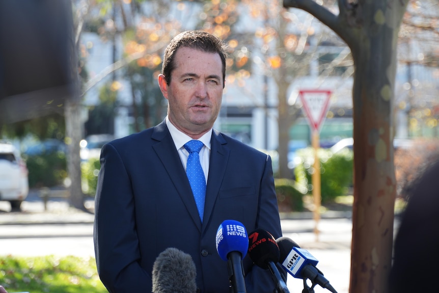 A middle-aged man with dark hair in a dark blue suit, white shirt and blue tie stands outside speaking at a media conference.