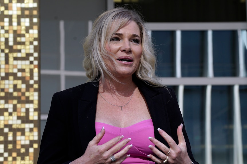 A woman with blonde hair wearing a black blazer speaks with her hands.