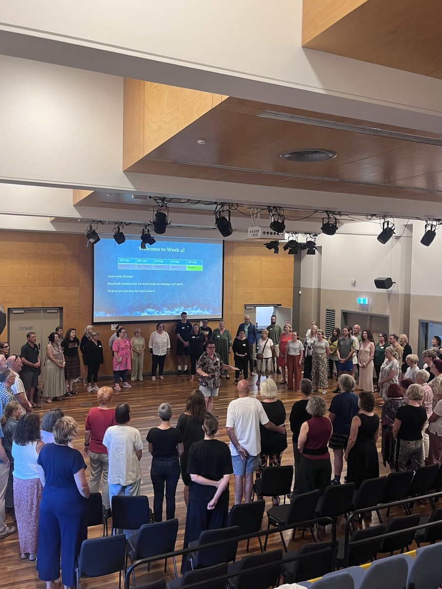 A man stands in the middle of a circle inside a community hall, surrounded by people as he teaches them a song.
