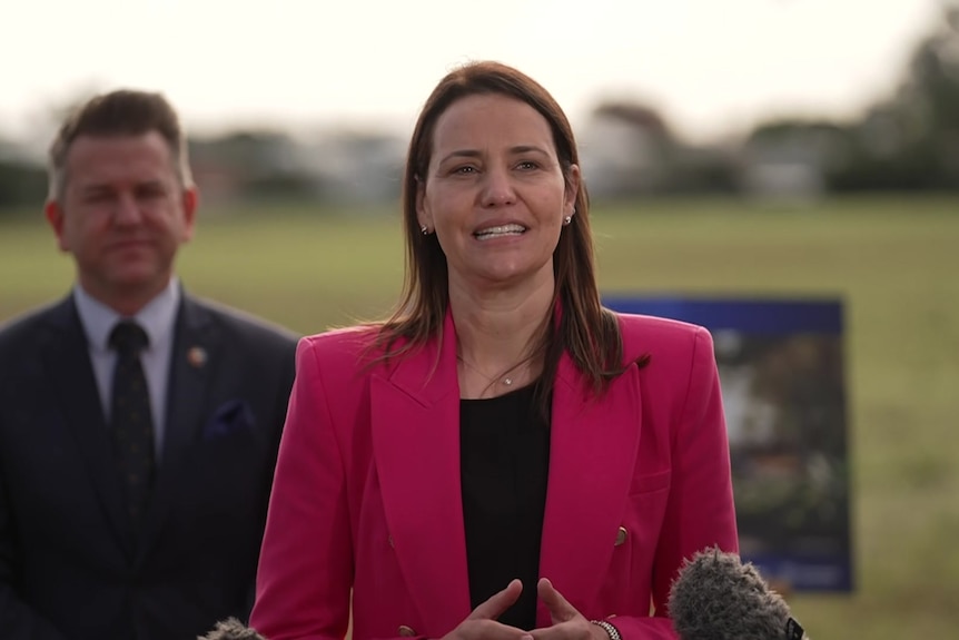 A woman in a pink blazer speaking into microphone with man in suit behind her.