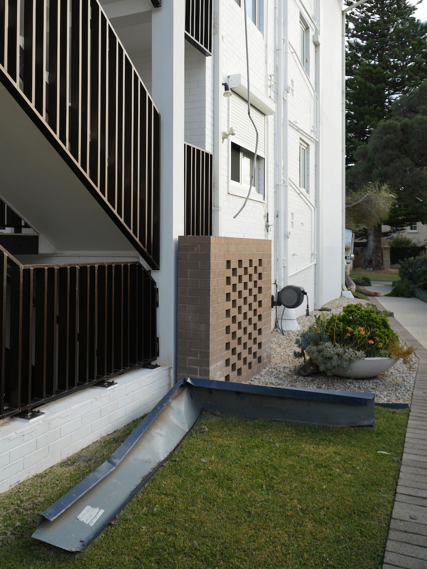 Part of a gutter from a roof lies on grass next to a white apartment building.