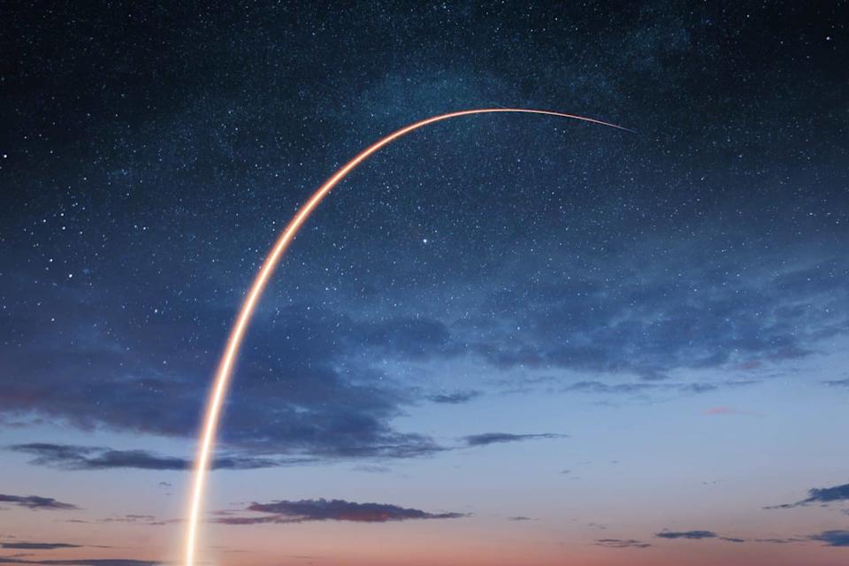 A long-exposure image of a rocket launch showing a bright, glowing orange light trail curving in an arc from the lower-center upwards into a deep blue night sky. Thousands of small white stars dot the dark sky above scattered, dark clouds, with the horizon showing faint hues of lighter blue and orange.