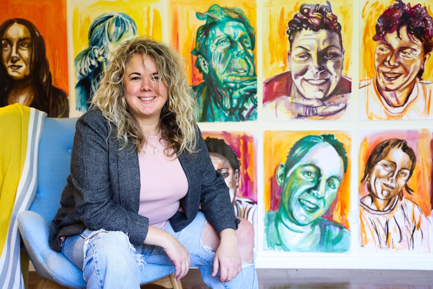 A woman with blonde curly hair looking at the camera. She is sitting on a blue chair in front of portraits of a group of women.