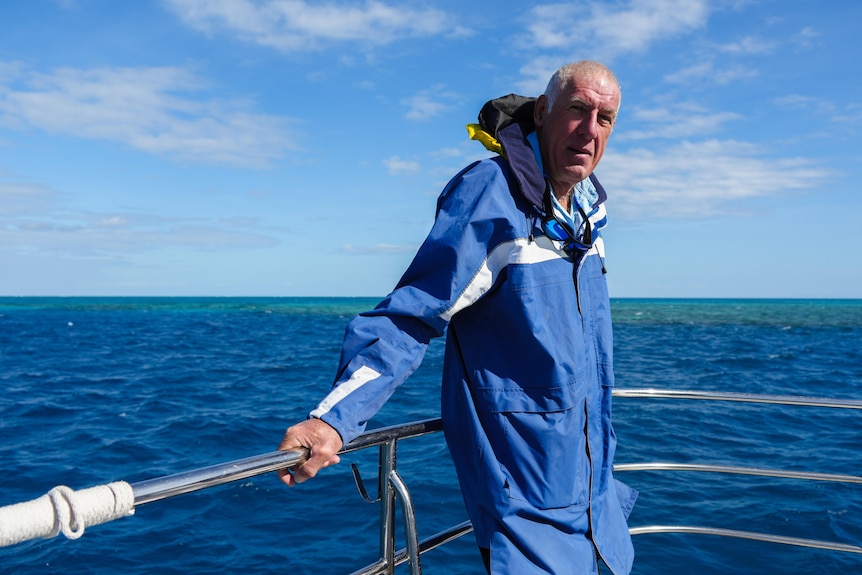 A man standing on the front of a boat holding onto the hand rail with water behind.