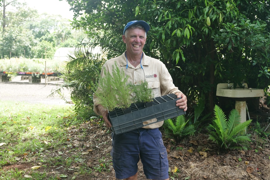 A man in outdoor workwear stands in front of a nursery, holding a tray of seedlings and laughing.