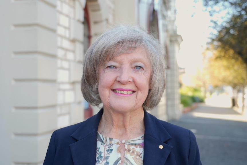 An older woman with silver-blonde hair wearing a blue blazer and a flowery top stands outside a historic white-painted building.