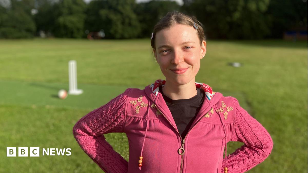 A woman wearing a pink zip up top. She is stood in a field with cricket stumps in the background.