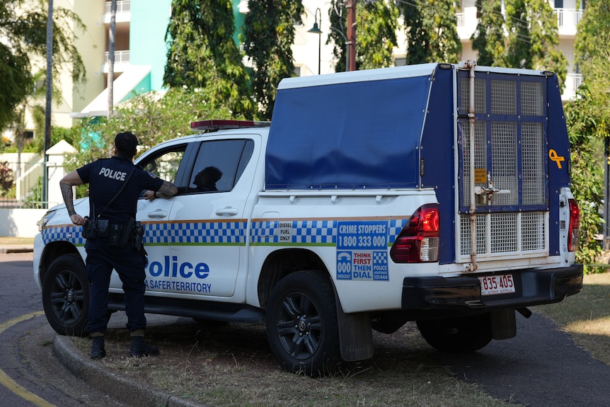 A uniformed police officer standing next to an NT Police caged vehicle, parked in a residential street.