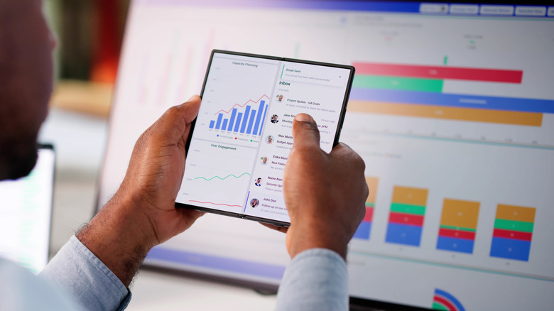 Person using a foldable phone to view business charts and messages in front of a large analytics display.