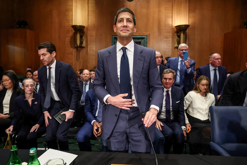 Kevin Warsh, standing smiling, with people in suits leaving their seats behind him.