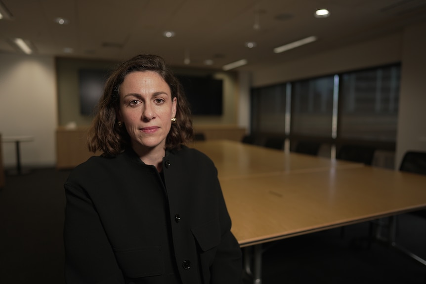 Carly Kind, wearing a black jacket, sits at a board room table.