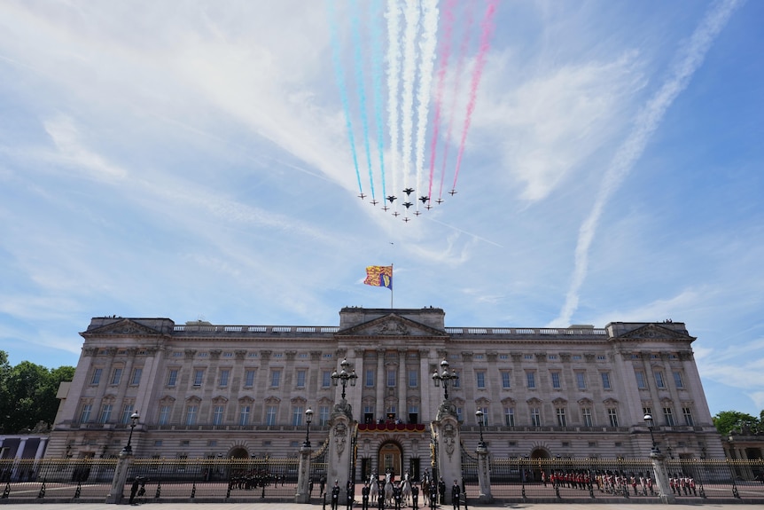 Jets fly over a palace in a clear blue sky leaving a trail of red, white and blue smoke behind them.