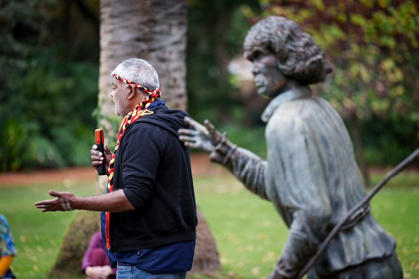 An Indigenous person stands next to the Balbuk Yooreel statue in a park