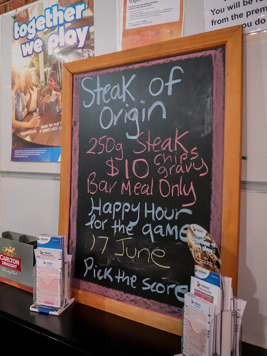 A blackboard propped on a table at a pub reads 'Steak of Origin' with price listed 'Happy Hour for the game'