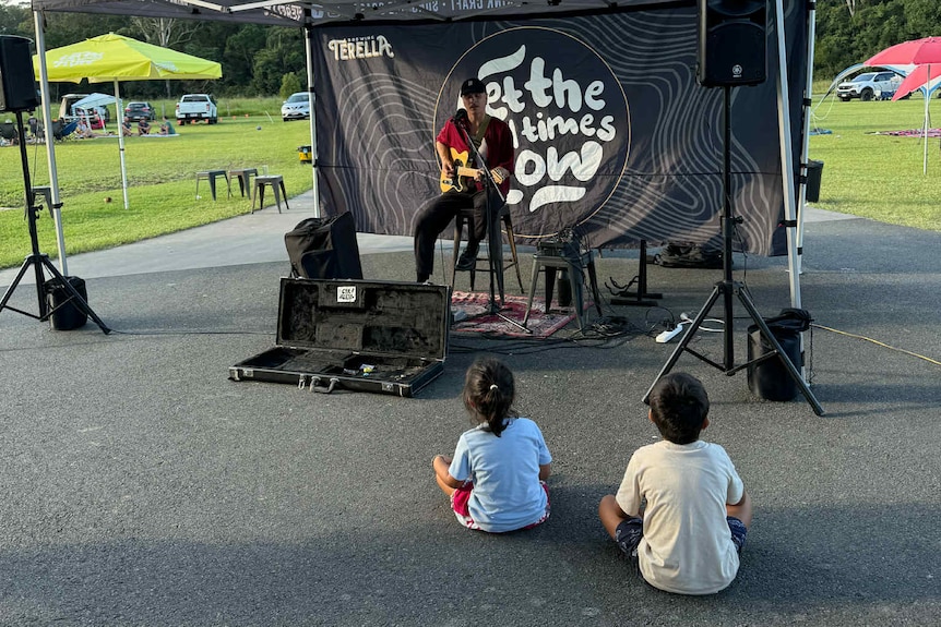 A quitarist performs under a tent as two children sit and listen