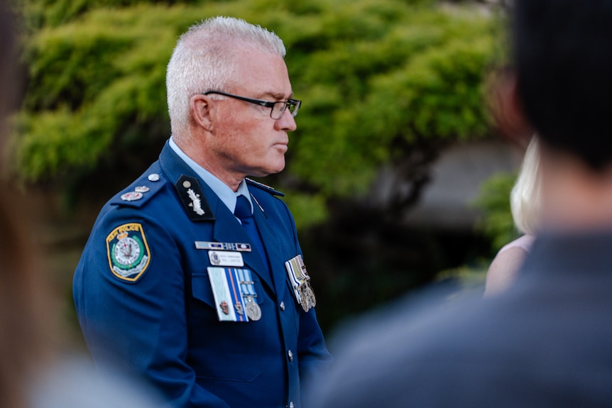 White main with cropped hair and glasses wearing police uniform with medals speaking to media.