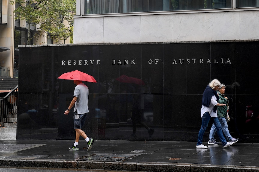 A man holding a red umbrella and two women walk past a black wall that reads Reserve Bank of Australia..