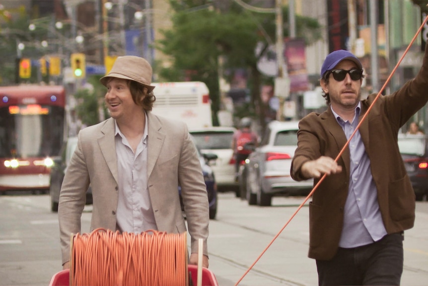 Two men on a busy city street unroll an orange electrical cable.