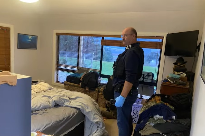 A police officer in a navy uniform with blue plastic gloves on his hands stands side-on in a bedroom of a house.