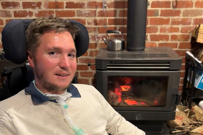 A smiling man in a wheelchair with a tracheostomy tube next to a fireplace