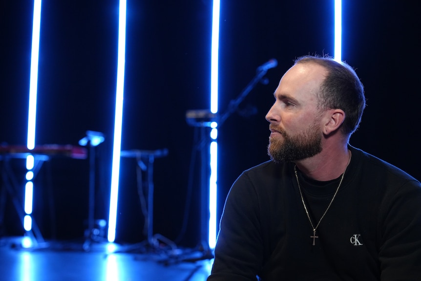 A side profile of Ryan who sits on a stage illuminated by blue light