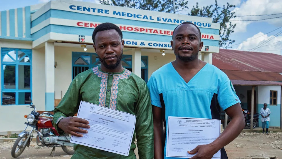 Tagirabo is wearing green and Bulambula is wearing turquoise. they are each holding a white certificate they received for recovering, and are standing outside a medical centre