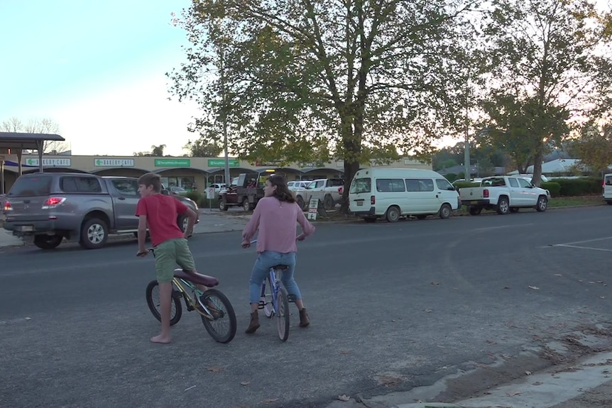 Two children on bikes wait for traffic to pass on a street.