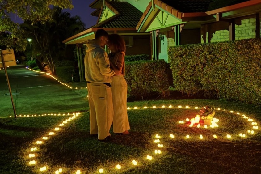 A young man and a young woman embrace on a lawn, which is surrounded by tealight candles in a love heart shape.