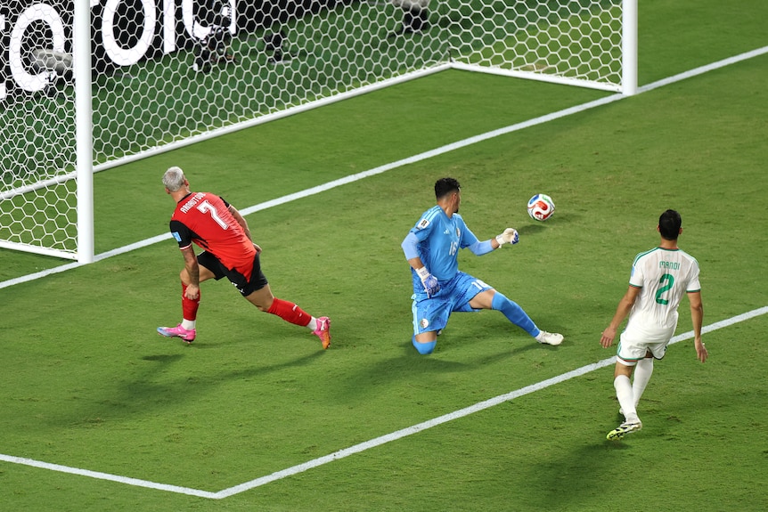 A soccer player in red rolls a ball into the goal with a goalkeeper beside him