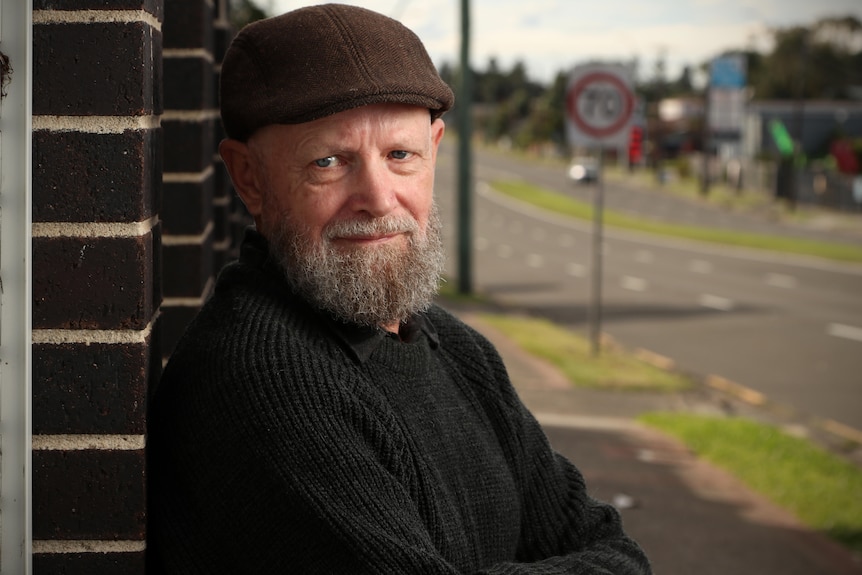 A bearded, middle-aged man in a flat cap leans against a brick wall near a main road.