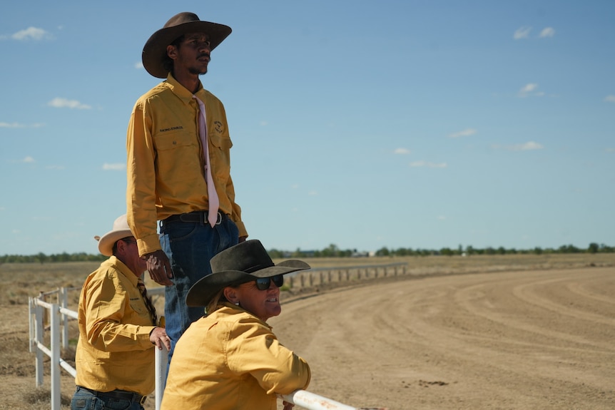 Man in cowboy hat stands on a fence to watch something ahead on a desert horse racing track 