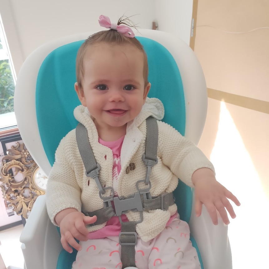 A baby wearing a pink hair tie sits in a blue and white high seat smiling at the camera